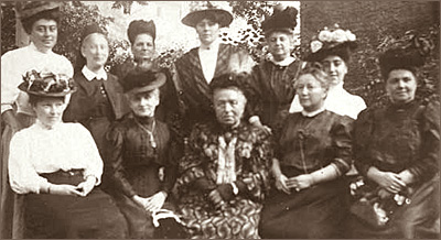 Foto av sex stående kvinnor och fem sittande. Det ser ut att vara utomhus och alla har stora hattar och gammaldags kläder