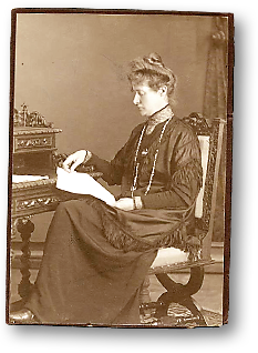 Foto av Lida Gustava Heyman som sitter på en utsirad stol vid ett utsirat, tjusigt skrivbord, och läser en tidnign eller ett dokument av något slag