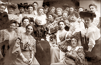 Foto av en stor grupp kvinnor som sitter och står iförda blusar och långa kjolar, en del med fina spetsar, och de flesta ser in kameran och ser glada ut