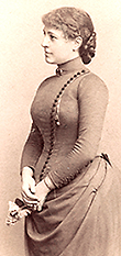 Foto i halvfigur och profil av Marit Stritt i unga år. Hon står rak, klänningen har knappar från axlarna och ner mot magen som utsmyckning och hon håller en blomma i handen