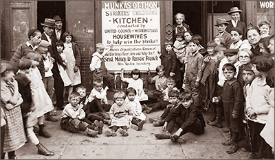 Foto av massor av barn och några vuxna som syåt på gatan och i dörröppningar. Mitt i bilden finns på väggen ett plakat där det står att det är "Strikers Childrens' Kitchen", ordnat av United Council of Workingclass Housewives "to help win the strike!". Det uppmanar också kvinnor och kvinnoorganisationer gå med och att stötta ekonomiskt