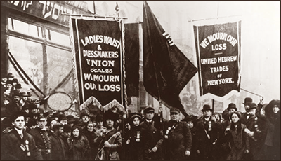 Foto av stor folksamling med stora standar. På ett står det "Ladies Waist & Dressmakers Union Local 25 We Mourn Our Loss" och på en annan står det "We mourn our loss - United Hebrew Trades of New York" De andra två går inte att se texten på. Alla ser allvarliga ut.