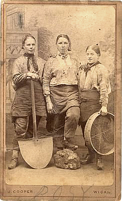 Tre byxklädda tjejer. En har en stor spade, en har en sorts sil och den tredje har foten på en stor sten