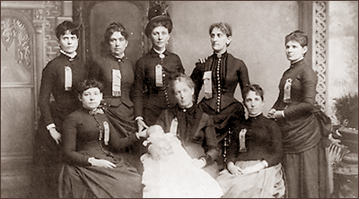 Foto av åtta kvinnor, fem stående och tre sittande, den i mitten har en baby i famnen, alla har långa klänningar