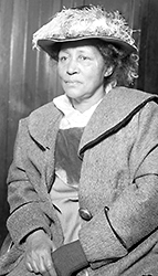 Foto av medelålders Lucy Parsons som sitter iförd hatt och kappa, med händerna i knät, och ser rakt framför sig