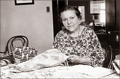 Foto av Marie Louise Bottineau Baldwin som sitter vid ett bord och håller i några textilier. Hon ser rakt in i kameran.