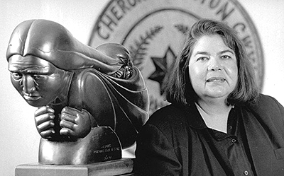 Foto av Wilma Mankiller bredvid en staty av en Cherokeekvinna med barn på ryggen. I bakgrunden syns symbolen för Cherokee Nation på väggen.