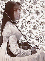 Foto i halvfigur av Zitkala-Ša som ung. Hon sitter i profil vänd åt höger, men har ansiktet riktat mot kameran. I knät har hon sin fiol och hon håller stråken upp mot axeln. På väggen bakom henne är en ljus tapet med stort blommönster