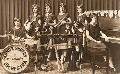 Foto av sex av systrarna i Schmitz Sisters Family Orchestra uppställda för fotografering med sina instrument. Viola sitter längst till vänster med sina trummor