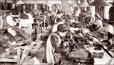 Foto från en sweatshop där ett stort antal kvinnor sitter och syr vid maskiner med tyg staplade runt omkring sig. I bakgrunden står några män och övervakar arbetet.