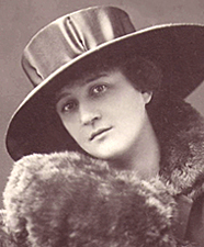 Porträttfoto av vacker ung Guilly med stor hatt och pälskrage. Hon ser allvarlig ut och tittar rakt in i kameran