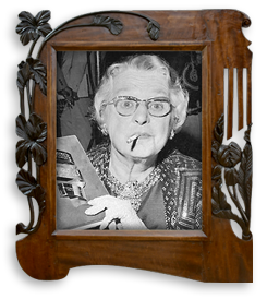 Svartvitt porträttfoto av Lotte Reiniger på äldre dar. Hon har glasögon på sig och har en cigarett i munnen. Ena handen syns och i den håller hon en bok eller liknande. Fotot är i en art nouveau -ram av mörkt trä med detaljer av växter i svart