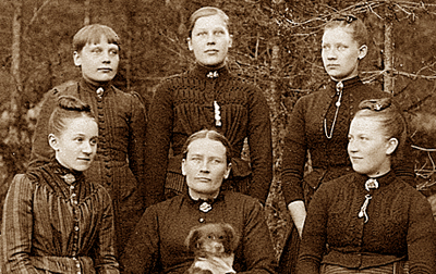 Foto av Karin Larsson sittande med hund i knät och omgiven av fem yngre kvinnor, varav åtminstone de tre bakersta hennes döttrar