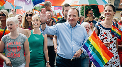 Annika Strandhäll och Stefan Löwén (S) går i Prideparaden, bägge vinkar till folk , Löwén håller en regnbågsflagga i vänstra handen.