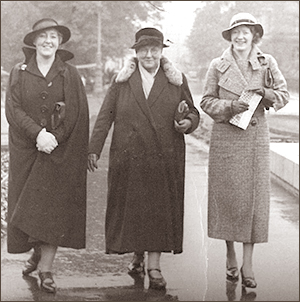 Foto av tre kvinnor i hattar och kappor som går i bredd på en gata