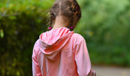 Foto av tjej i rosa tröja med huva, sett bakifrån. Hon har mörkt hår i två flätor med mörkrosa snoddar, bakgrunden är gröna buskar