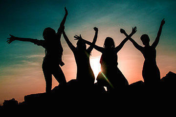 Foto av siluetterna av fyra tjejer som står med armarna upp i luften och solnedgång i bakgrunden